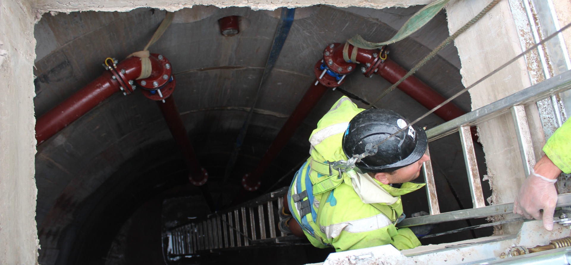 A T-T Engineer working on-site within a confined space, descending a ladder into a pumping station chamber below ground, wearing a hard hat, harnesses and other appropriate safety equipment.