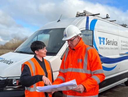 Photo of two people in orange high visibility jackets in front of a T-T Service van. One is a T-T Service engineer wearing a hard hat, looking at site plans with a cliant.