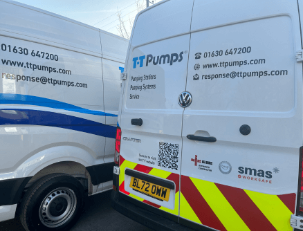 Photograph of the back of two white vans parked side by side, with the blue T-T Pumps logo, contact information, accreditatino logos and a QR code that leads to the website when scanned.
